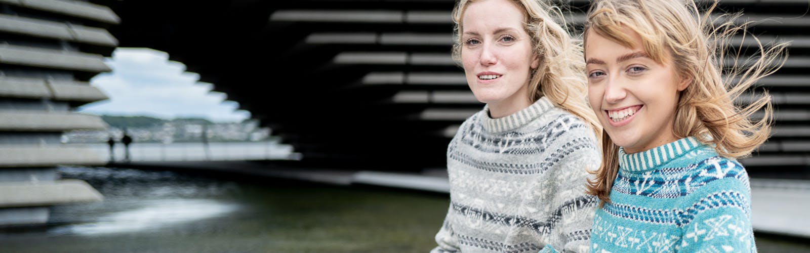 A photo of two women smiling and standing outside V&A Dundee wearing fair isle jumpers designed around jute, jam and journalism.